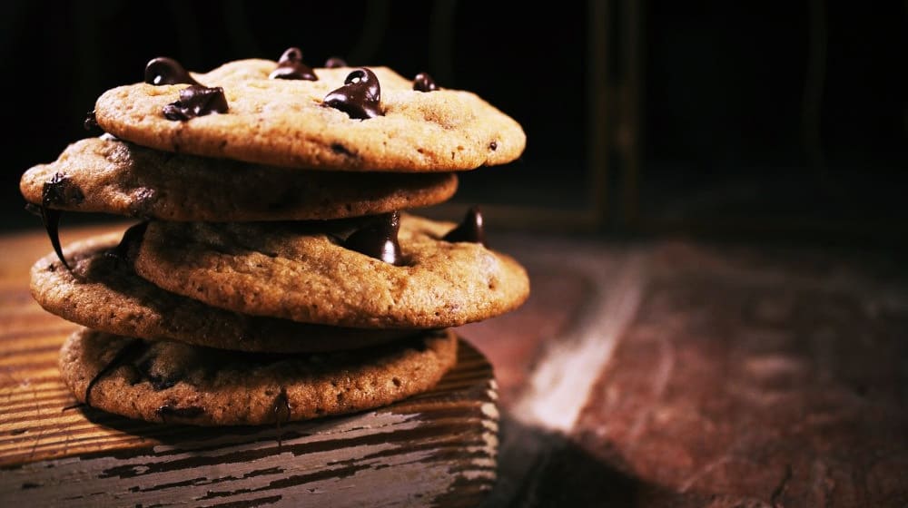 Ein Stapel Chocolate-Chip-Cookies auf einer Holzoberfläche, mit einem Hauch von Kratom, vor einem dunklen Hintergrund.
