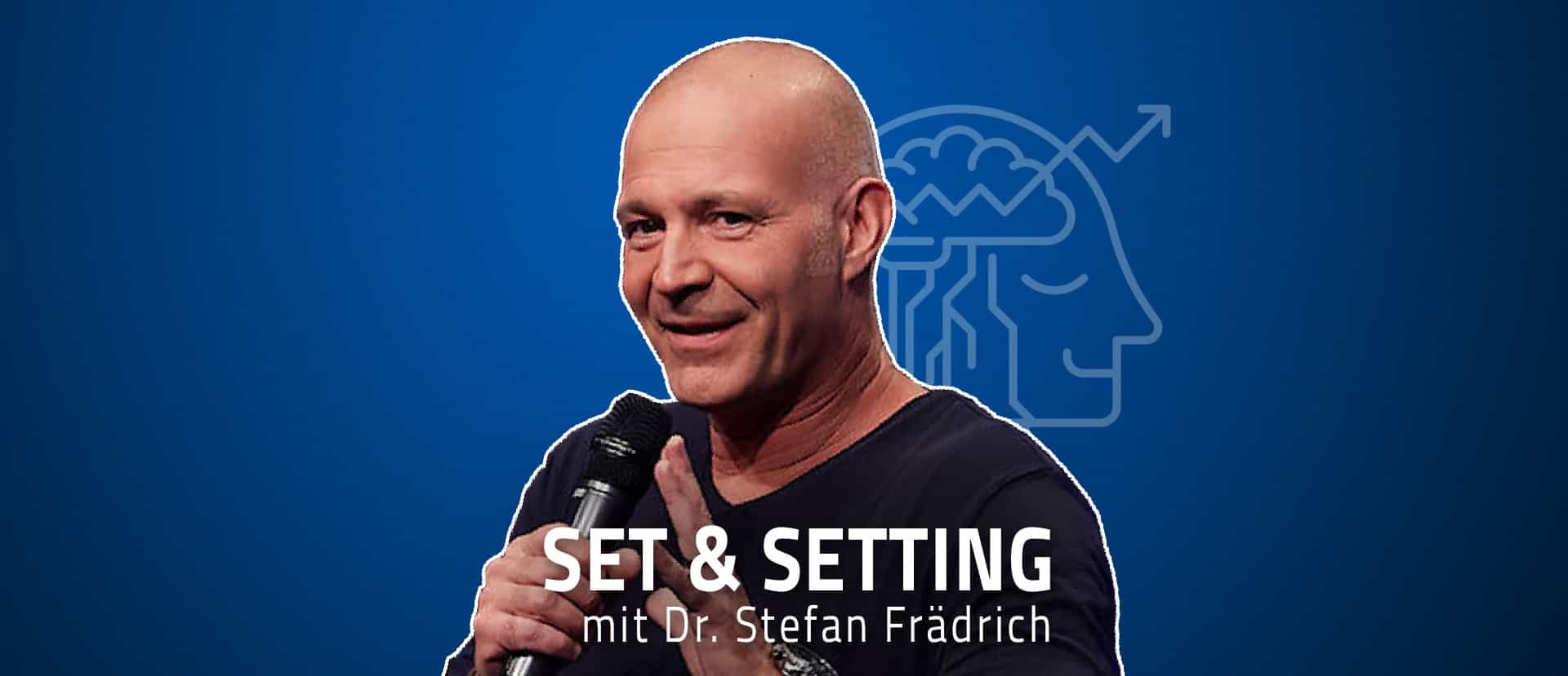 Ein kahlköpfiger Mann in einem schwarzen Hemd mit einem Mikrofon spricht auf der Bühne vor einem blauen Hintergrund, auf dem eine Gehirnillustration zu sehen ist, und thematisiert gesellschaftliche Probleme.