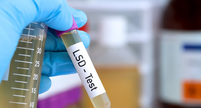 Eine behandschuhte Hand hält ein Reagenzglas mit der Aufschrift „LSD – Test“ in einer Laborumgebung mit unscharfer Ausrüstung im Hintergrund.